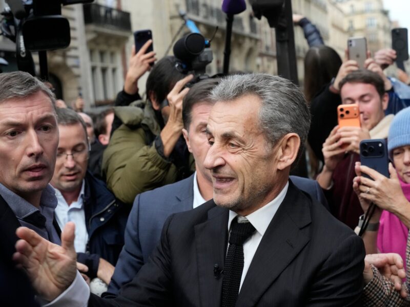 Nicolas Sarkozy smiles in a suit and tie amidst a crowd taking photos of him on a street.
