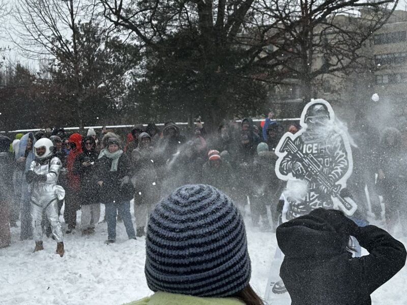 A circle of people dressed in heavy winter gear throw snowballs at a cardboard cutout of an ICE agent amid several inches of snow.