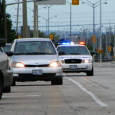 A police cruiser with flashing lights follows a car during a traffic stop on a multi-lane road.