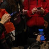 A group of girls stand with phones in hand around a girl sitting down, looking at her screen, on the metro.