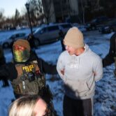 Two people with covered faces and tactical vests, one marked "Police ICE," hold a young man's arms behind his back as they stand in light snow.