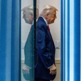 Donald Trump and his reflection seen as he walks past a glass door and blue curtain.