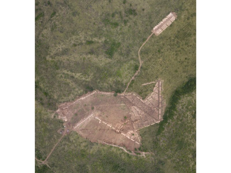 Aerial view of a cleared excavation site with grid-like digging patterns connected by dirt paths in a grassy landscape.