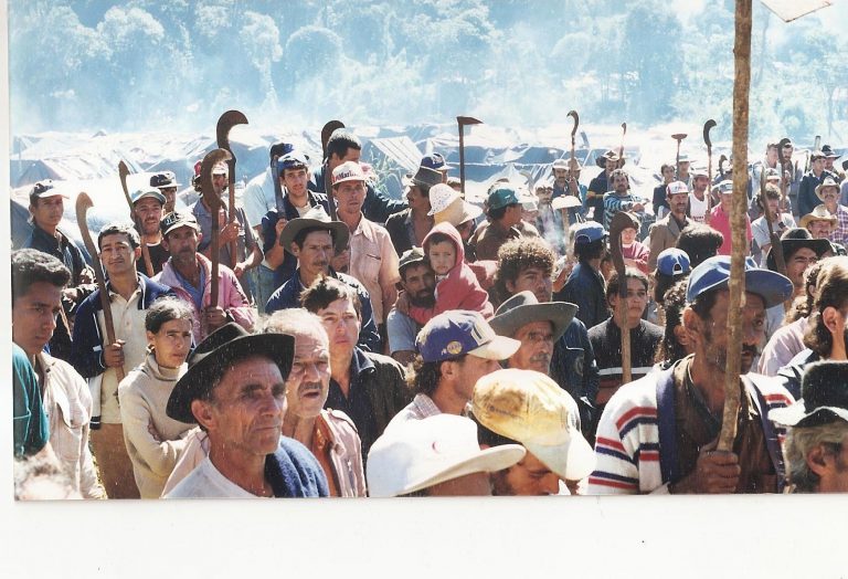 A crowd of people holding tools and wearing hats stands closely together in an outdoor encampment.