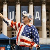 A man wearing a stars-and-stripes outfit points while standing in front of large “USA” banners.