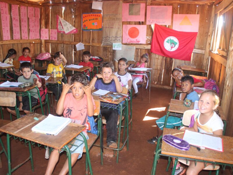 Children sit at desks inside a simple wooden classroom with posters on the walls.