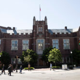 A large red-brick academic building on the campus of University of Pennsylvania stands at a street corner in Philadelphia as pedestrians cross the intersection.