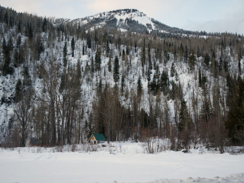 A snowy mountainside with scattered trees overlooking a frozen lake and a small cabin.