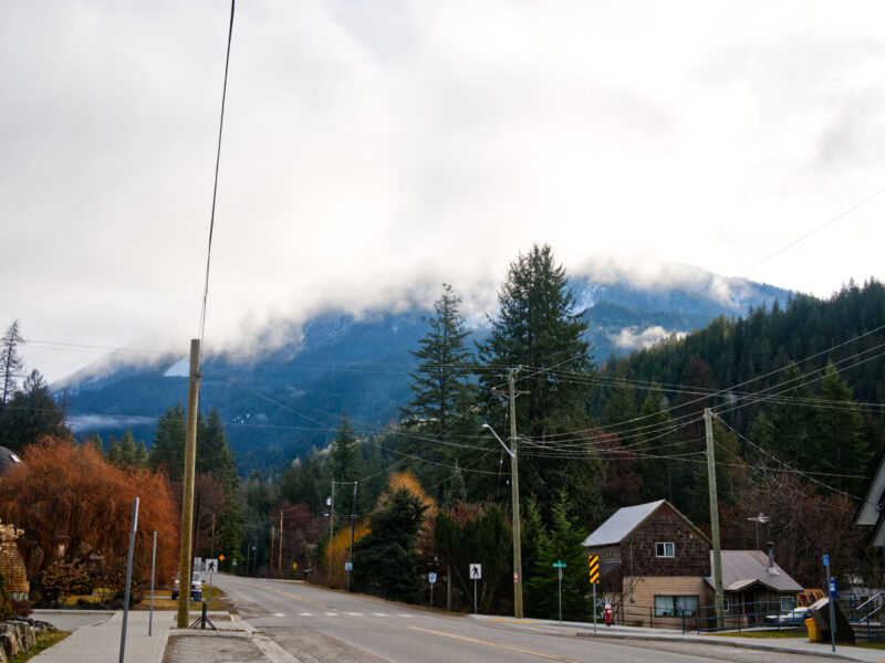 A small town street with houses and trees set against forested mountains partly covered in clouds.