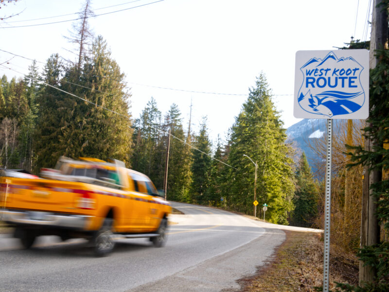 A pickup truck drives along a forested mountain road beside a roadside sign.