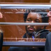 A frowning Black man sits behind a plastic shield, flanked by two women.