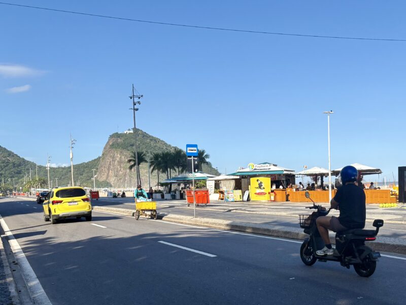 A person rides a small electric two-wheeler on a roadway next to the beach in Rio de Janeiro.