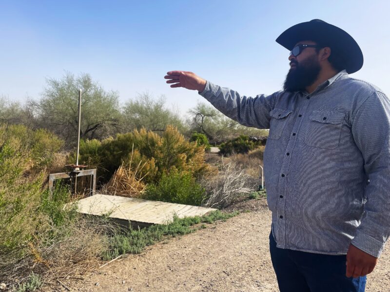 A man in a cowboy hat gestures toward brush and wetlands beside a dirt path.