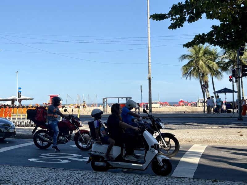 A woman and a child ride together on a white electric two-wheeler near the beach in Rio de Janeiro.