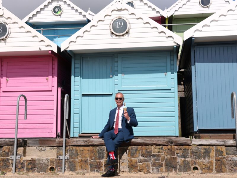 Nigel Farage, in Union Jack socks, a suit and sunglasses, holds an ice cream in front of pastel numbered bungalows.