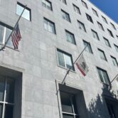 A large grey building with rectangular windows and the United States and State of California Flags hanging off.