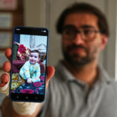 Fahd Abu Haikal holds up a cellphone displaying a photo of his infant son while standing indoors with his hand wrapped in a bandage in Hebron, West Bank.