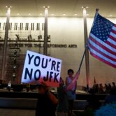 A small group of protesters gathers outside the John F. Kennedy Center for the Performing Arts in Washington as crews and scaffolding are visible along the building's facade.