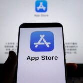 The App Store blue and white "A" logo on a phone, in front of a computer screen with the same logo.