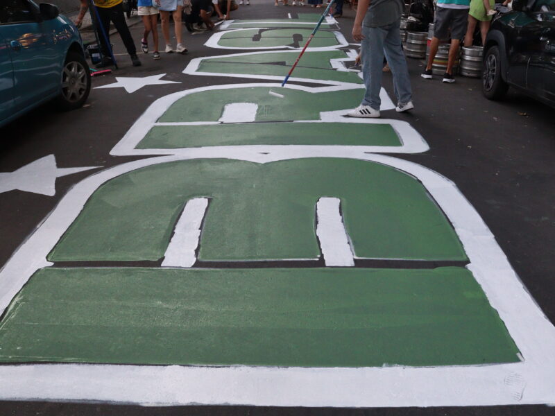 Artists use paint rollers to fill in large green letters spelling 
