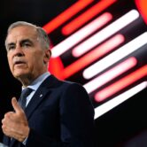 Canadian Prime Minister Mark Carney speaks at a podium during a defense industry conference in Ottawa, gesturing with his hand as red and white illuminated panels glow in the background.