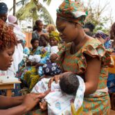 A woman holds a baby wrapped in a blanket as another woman wipes the baby's arm.