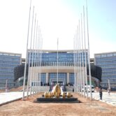 A multistory building with two symmetrical wings and an entryway lined with two rows of tall poles.