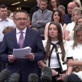 A man and two women stand side by side as he reads from a sheaf of papers into a microphone.