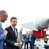 Friedrich Merz and René Obermann talk in front of three people in suits and military dress under a plane.