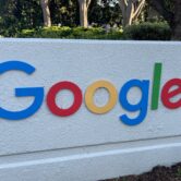 Google's multicolor logo on a cream colored sign with trees in the background.