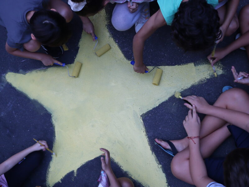 Children sit around a large yellow star painted on the pavement, using brushes and rollers during a World Cup street-painting event.