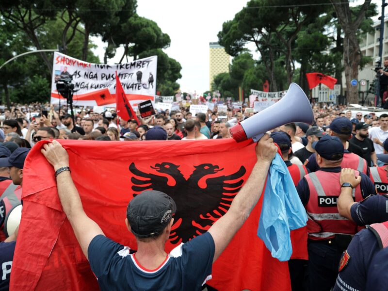 A large crowd gathers in central Tirana during a protest against a proposed resort development, as a demonstrator in the foreground raises an Albanian flag while police officers stand nearby.