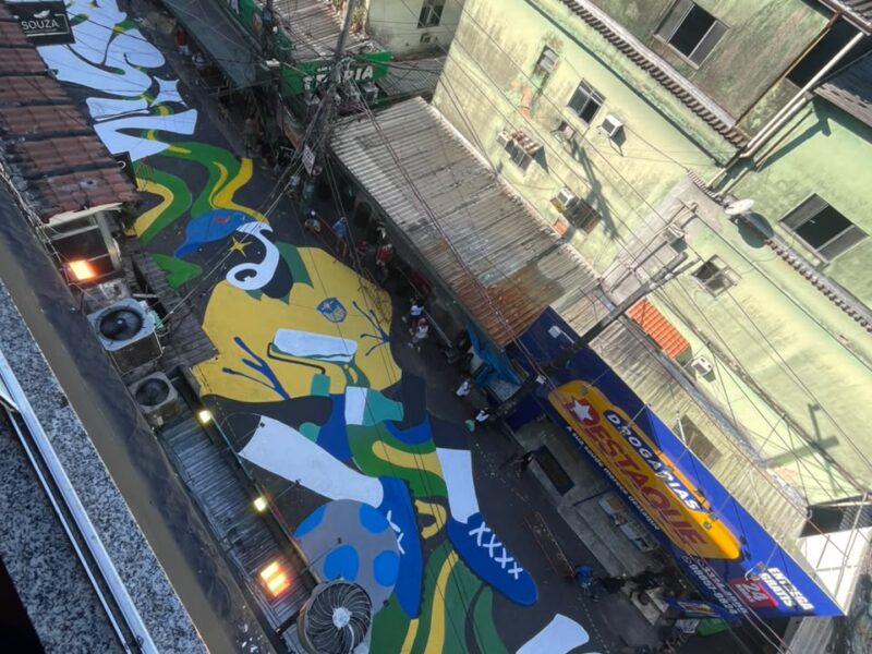 Aerial view of Via Apia in Rocinha, with a large green, yellow and blue World Cup mural covering the narrow street between buildings.