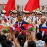 Xi Jinping and Vladimir Putin are seen with officers in dress uniform holding bayonets behind them and children facing them.