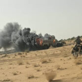 Black smoke and flames rise from burning military vehicles in a desert landscape as a motorcyclist rides nearby.