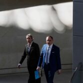 A blond woman and a balding man walk in a shadow-filled hallway.