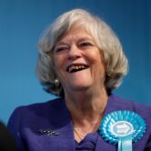 A woman with a silver bob smiles against the blue sky. She's wearing a royal blue jacket with a blue ribbon pinned on the lapel.