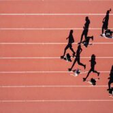 Aerial view of athletes running in separate lanes on a red track, casting long shadows.