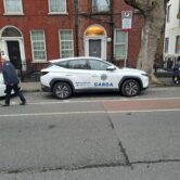 A Garda police SUV parked on a street outside red-brick buildings, with pedestrians walking past and a group gathered on the sidewalk.
