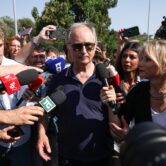 A man with gray hair, in sunglasses, is surrounded by reporters with microphones and cameras.