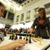 A woman stands as she plays a young person at chess as people sit with chessboards around a long table.