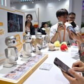 People take pictures at a trade booth with small silver robots displayed on a table.