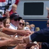 Trump shakes hands with a crowd of people across a barrier with a train in the background.