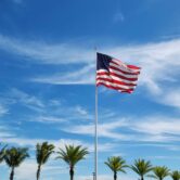 A U.S. flag flies high above a row of palm trees, against a blue sky with swirls of white clouds.