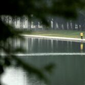Two workers in reflective yellow and orange vests walk along the reflecting pool from a distance, framed by tree limbs.